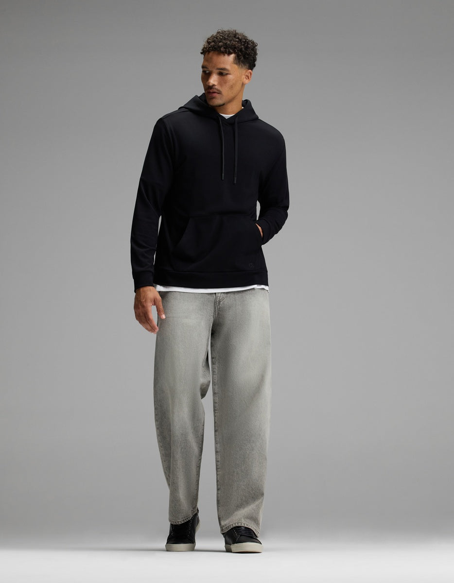 A man stands against a plain gray background, looking to his left with one hand in the pocket of his Cozy Earth Men’s Brushed Bamboo Hoodie, layered over a white shirt and paired with loose-fitting gray jeans.