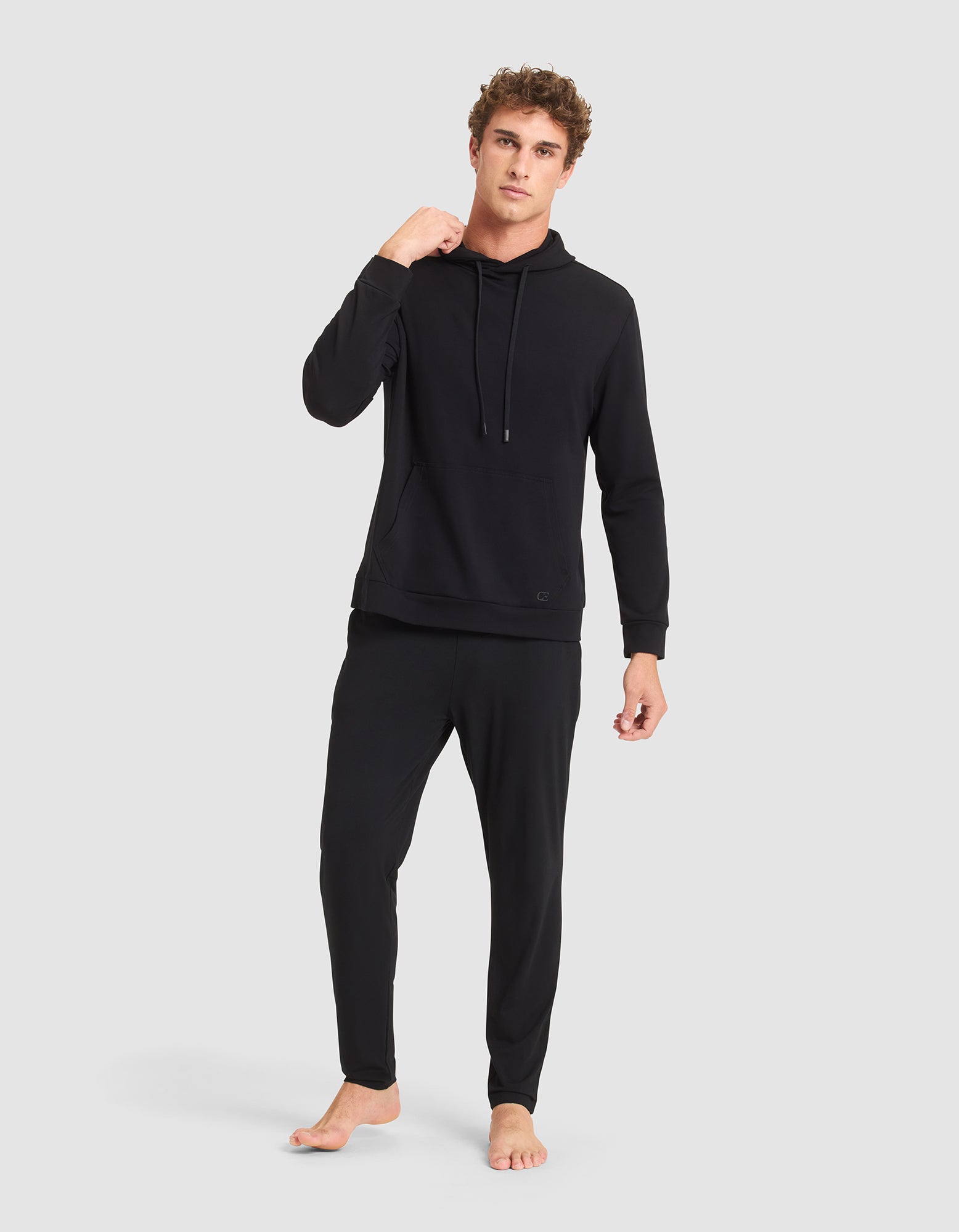 A barefoot man with curly hair stands against a light gray background, wearing Cozy Earth's Men's Brushed Bamboo Hoodie and matching jogger pants. He has a relaxed expression. 