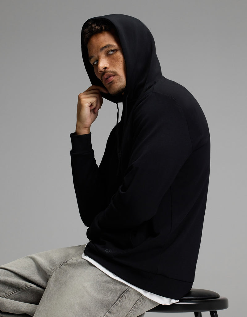 A man wearing the Cozy Earth Men’s Brushed Bamboo Hoodie and gray jeans sits on a stool against a plain gray background, looking seriously at the camera with one hand near his chin.