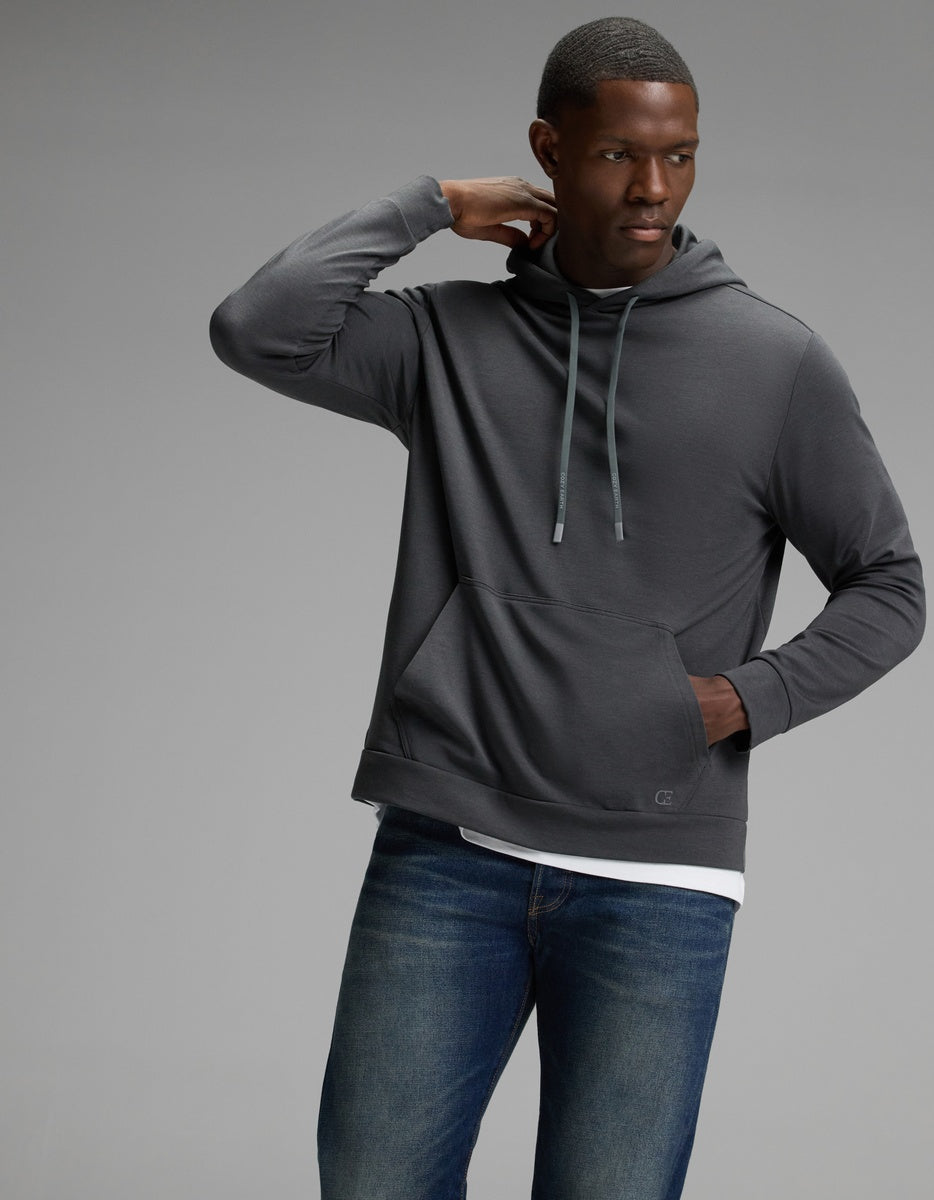 A man models the Cozy Earth Men’s Brushed Bamboo Hoodie in dark gray with blue jeans, standing against a plain gray background and gazing to the side while resting one hand on the back of his neck.