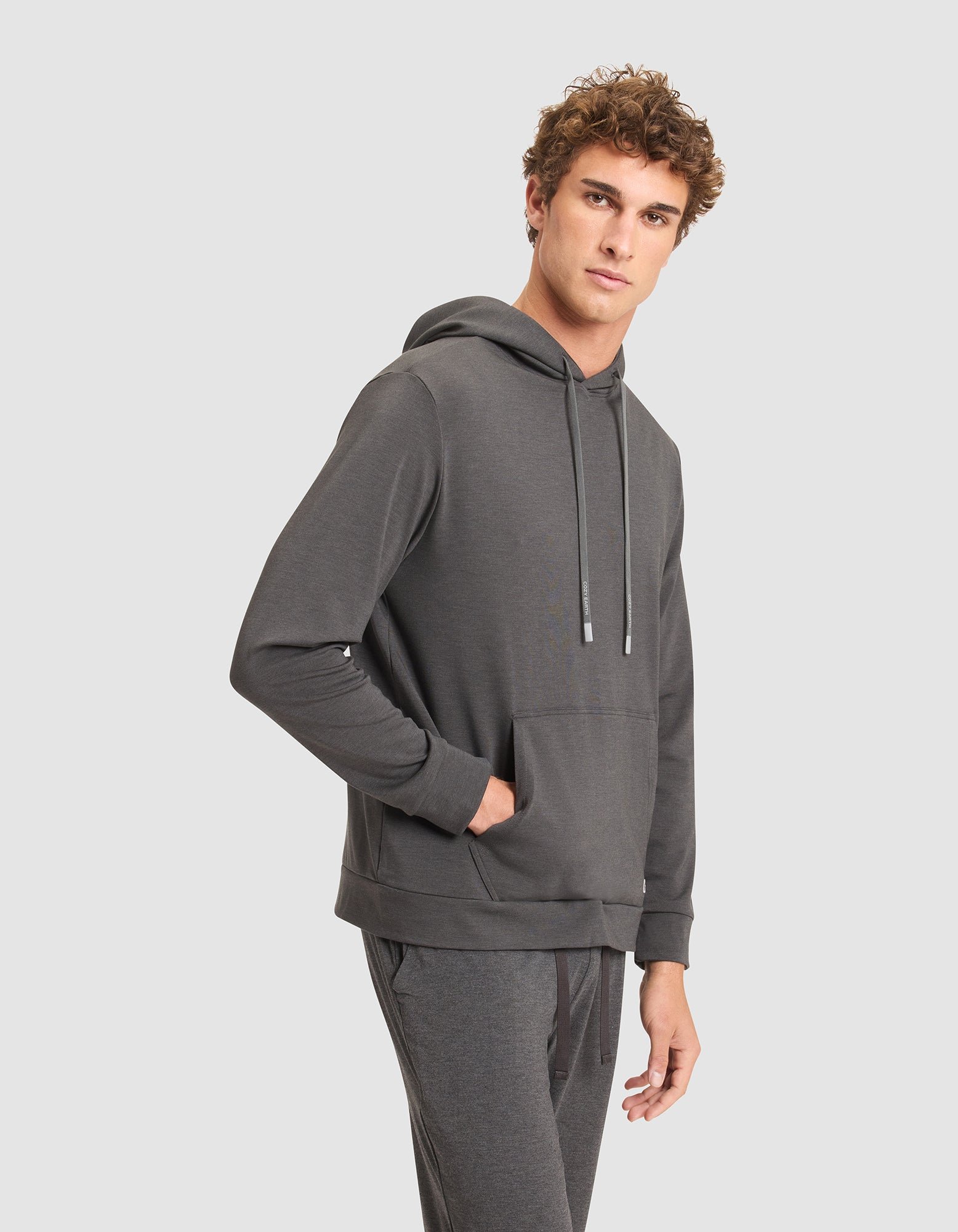 A young man with curly hair models the Cozy Earth Men's Brushed Bamboo Hoodie in dark gray, paired with matching joggers, standing with one hand in his pocket against a plain light gray background. 