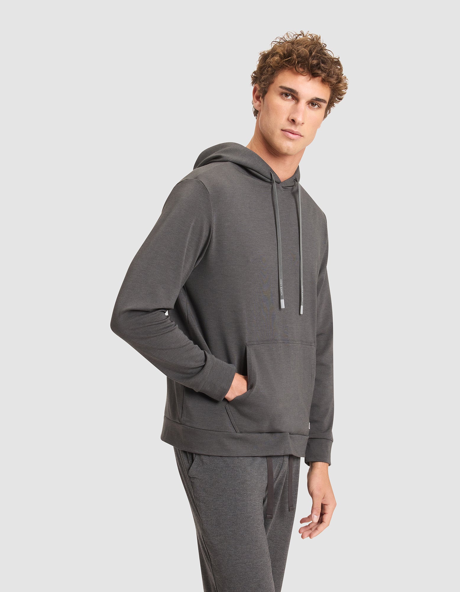 A young man with curly hair models the Cozy Earth Men's Brushed Bamboo Hoodie in dark gray, paired with matching joggers, standing with one hand in his pocket against a plain light gray background. 