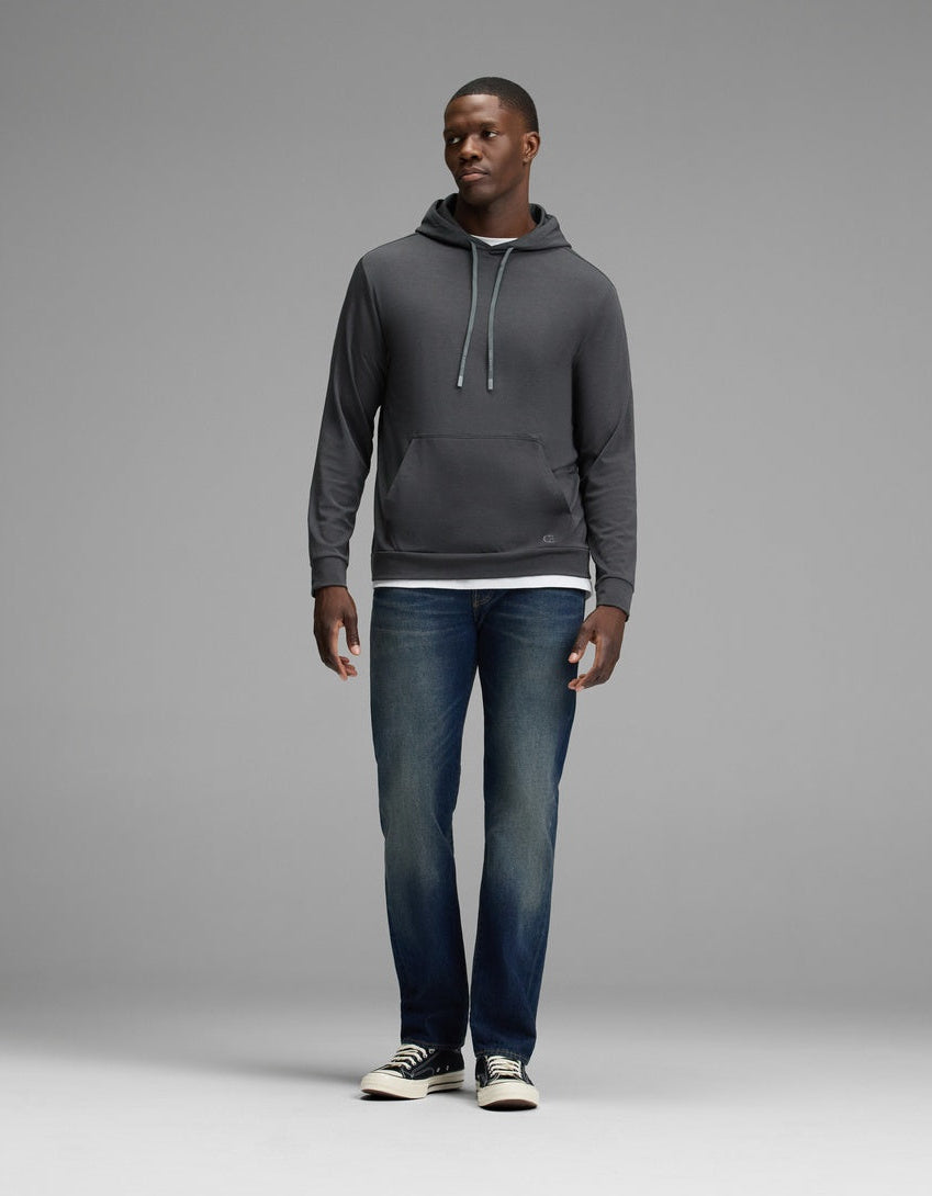 A man stands against a plain gray background wearing the Cozy Earth Men’s Brushed Bamboo Hoodie in dark gray, paired with blue jeans and black-and-white sneakers. He faces slightly to the side with his hands relaxed by his sides.