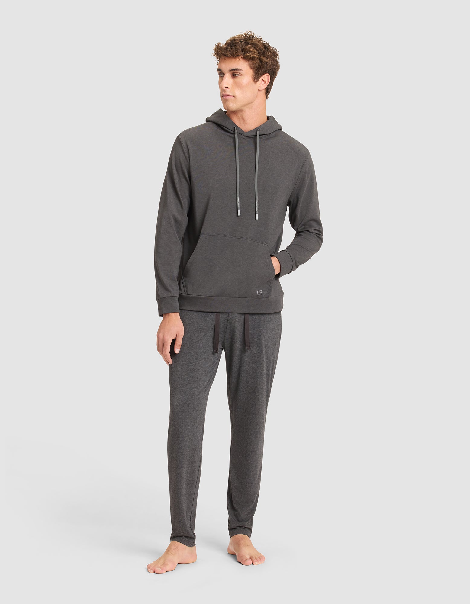 A man stands barefoot in the Cozy Earth Men's Brushed Bamboo Hoodie, a dark gray hooded sweatshirt with drawstrings and matching relaxed-fit pants, posing against a plain light background. 