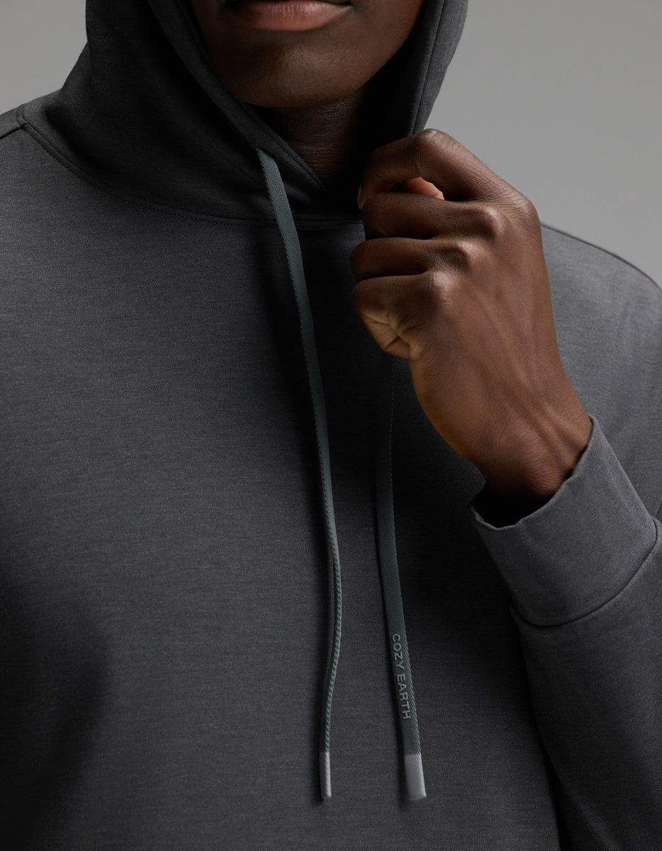 A person wearing the Cozy Earth Men’s Brushed Bamboo Hoodie in dark gray holds the hood’s drawstring—featuring a subtle logo—near their chin, with a plain gray background.