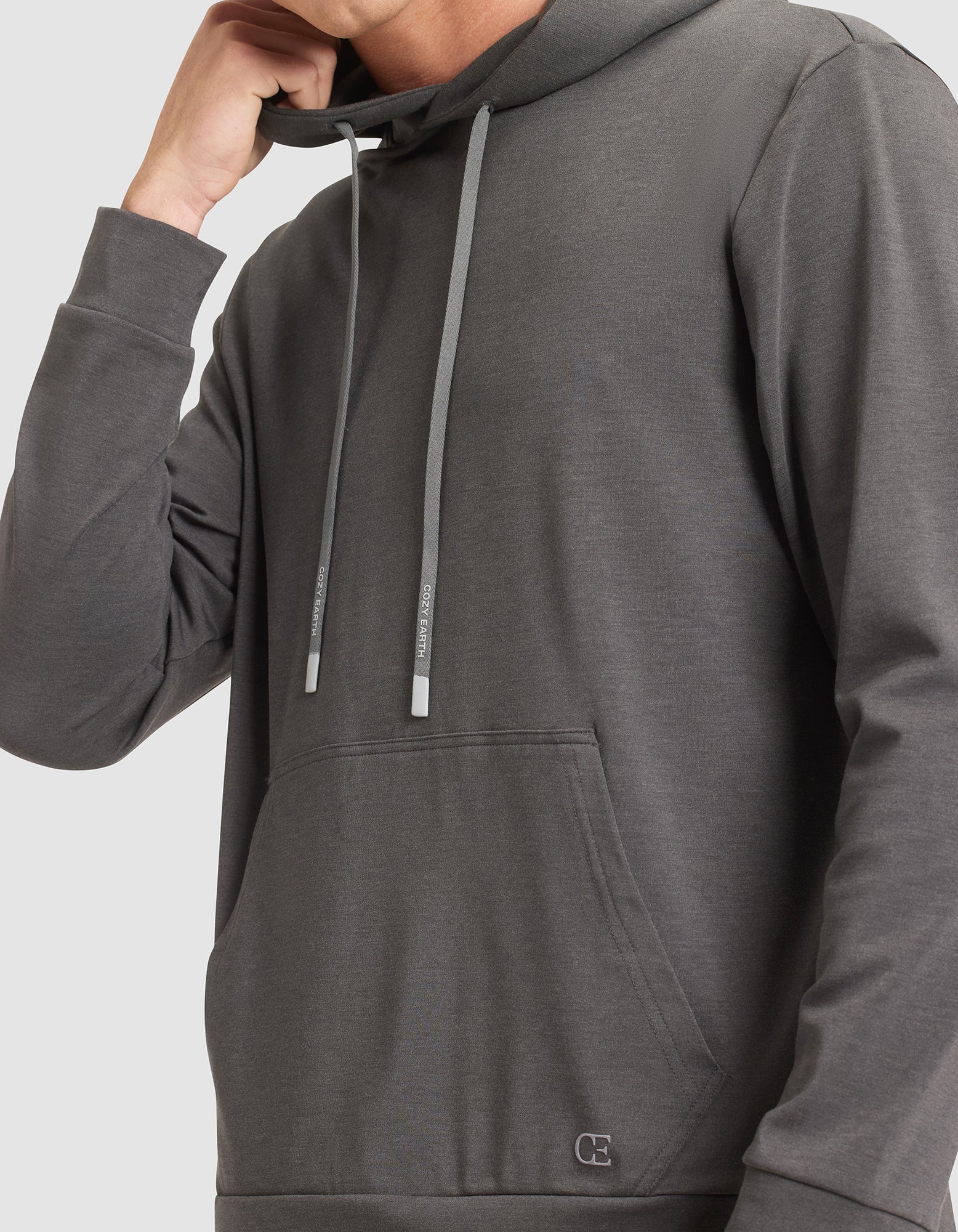 A person wearing the Cozy Earth Men's Brushed Bamboo Hoodie in dark gray, featuring a front pocket and drawstrings, is shown from shoulders to waist with their left hand raised near their neck. 