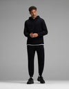 A man stands against a gray background, wearing the Cozy Earth Men’s Brushed Bamboo Hoodie & Jogger Set in black, with a white T-shirt visible beneath the hoodie and black sneakers. He looks slightly to the side, hands together.|Color:Black