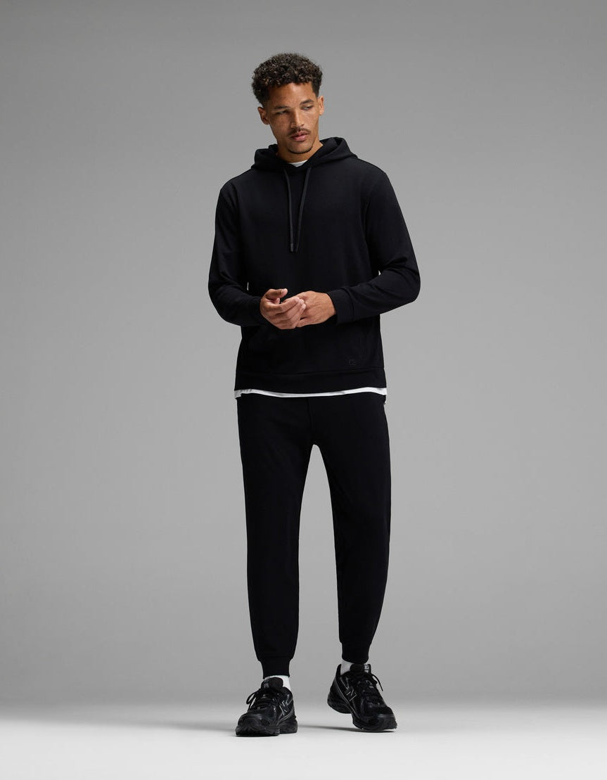 A man stands against a gray background, wearing the Cozy Earth Men’s Brushed Bamboo Hoodie & Jogger Set in black, with a white T-shirt visible beneath the hoodie and black sneakers. He looks slightly to the side, hands together.