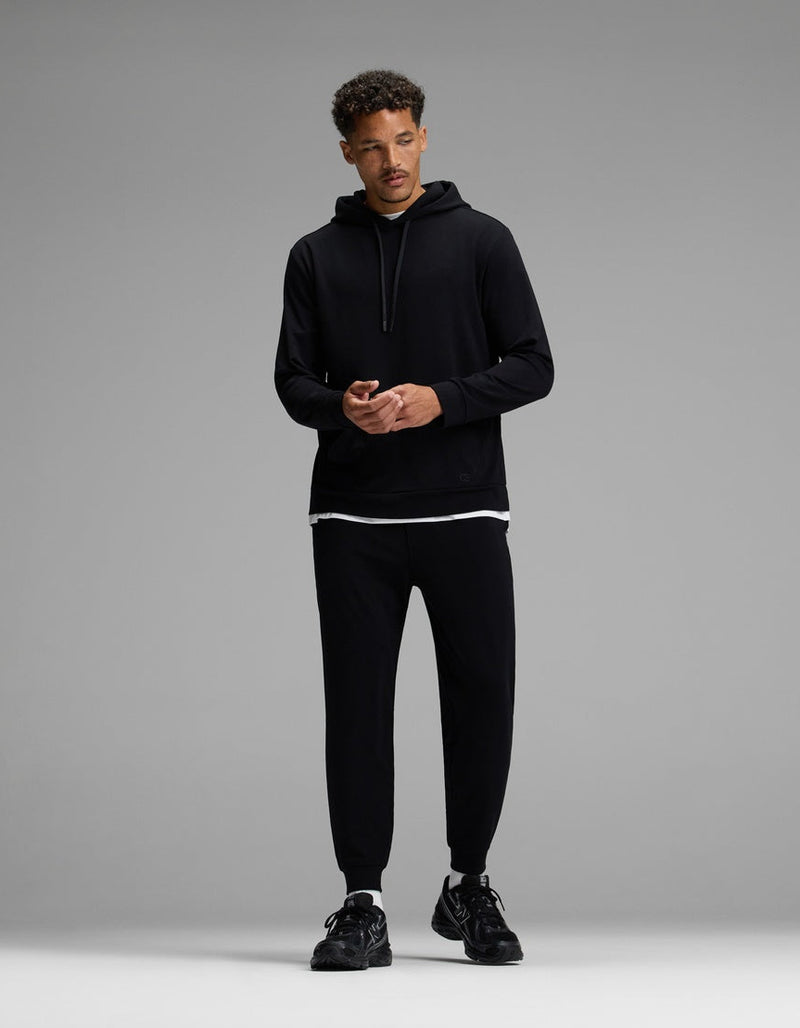 A man stands against a gray background, wearing the Cozy Earth Men’s Brushed Bamboo Hoodie & Jogger Set in black, with a white T-shirt visible beneath the hoodie and black sneakers. He looks slightly to the side, hands together.