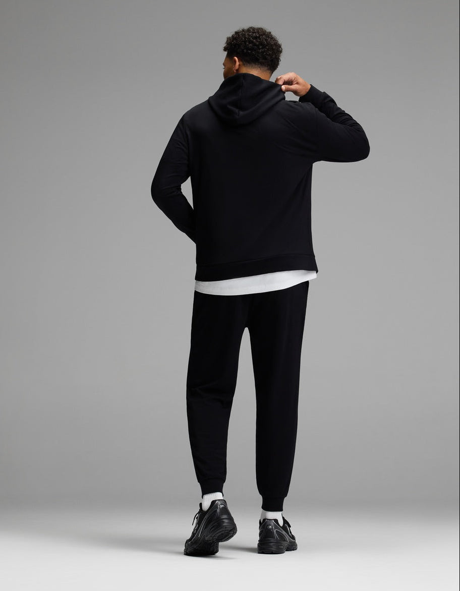 A person stands facing away, dressed in the Cozy Earth Men’s Brushed Bamboo Hoodie & Jogger Set in black, paired with black sneakers and a white shirt peeking out underneath. The background is plain gray.