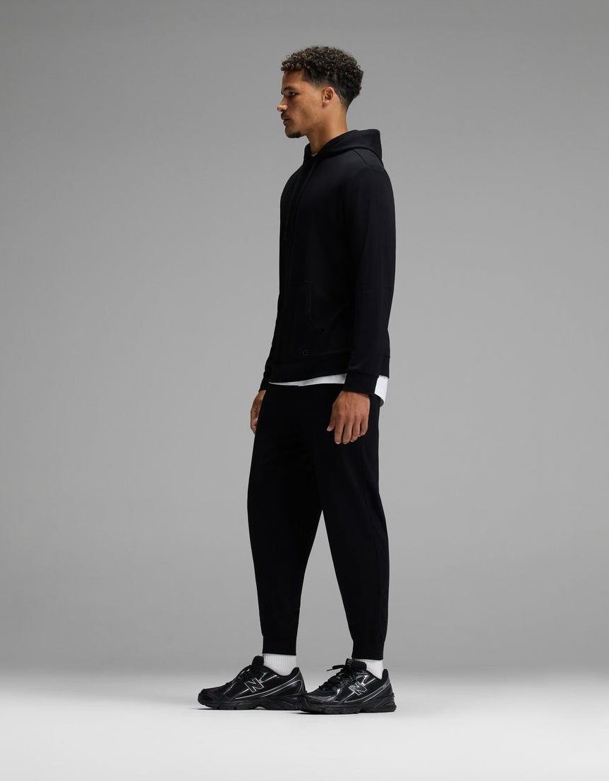 A man stands in profile against a plain gray background, wearing the Cozy Earth Men’s Brushed Bamboo Hoodie & Jogger Set in black, paired with black sneakers and a white shirt peeking out from under the hoodie.