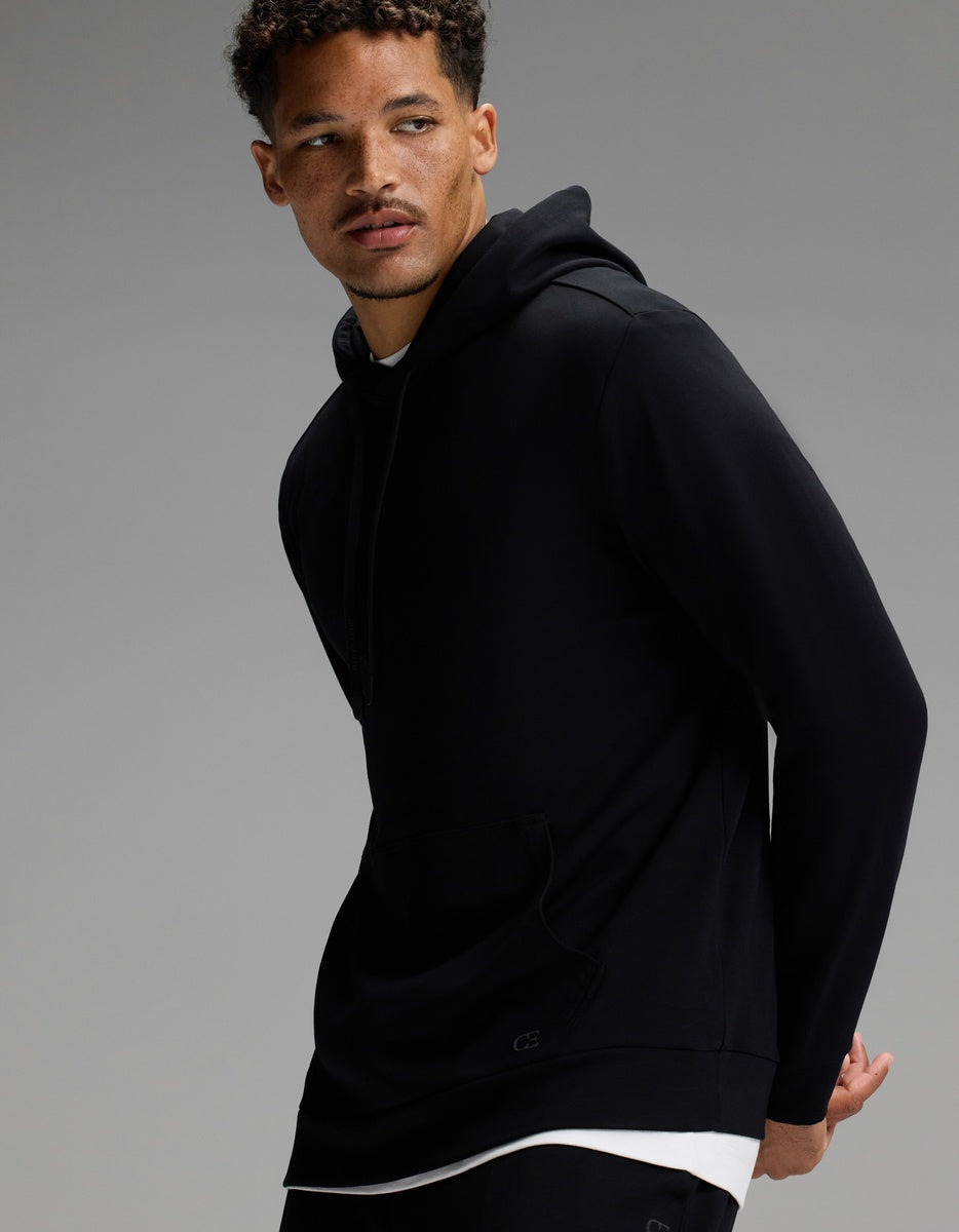 A man with short curly hair models the Cozy Earth Men’s Brushed Bamboo Hoodie & Jogger Set in black over a white shirt, posing against a plain gray background.