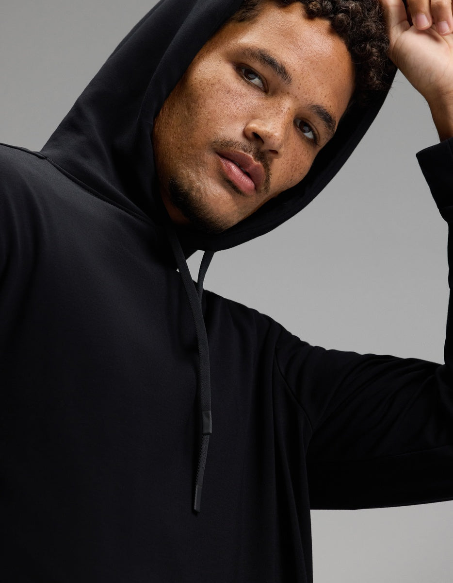 A person with curly hair and facial stubble, wearing the Cozy Earth Men’s Brushed Bamboo Hoodie & Jogger Set in black, looks at the camera with a neutral expression. Their left hand lifts the hood slightly, and the background is plain and light gray.