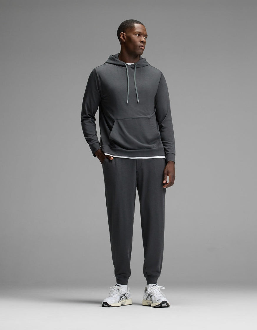 A man stands against a grey background, wearing the Cozy Earth Men’s Brushed Bamboo Hoodie & Jogger Set in Charcoal, paired with white athletic shoes. He looks to the side with his hands by his sides.|Color:Charcoal
