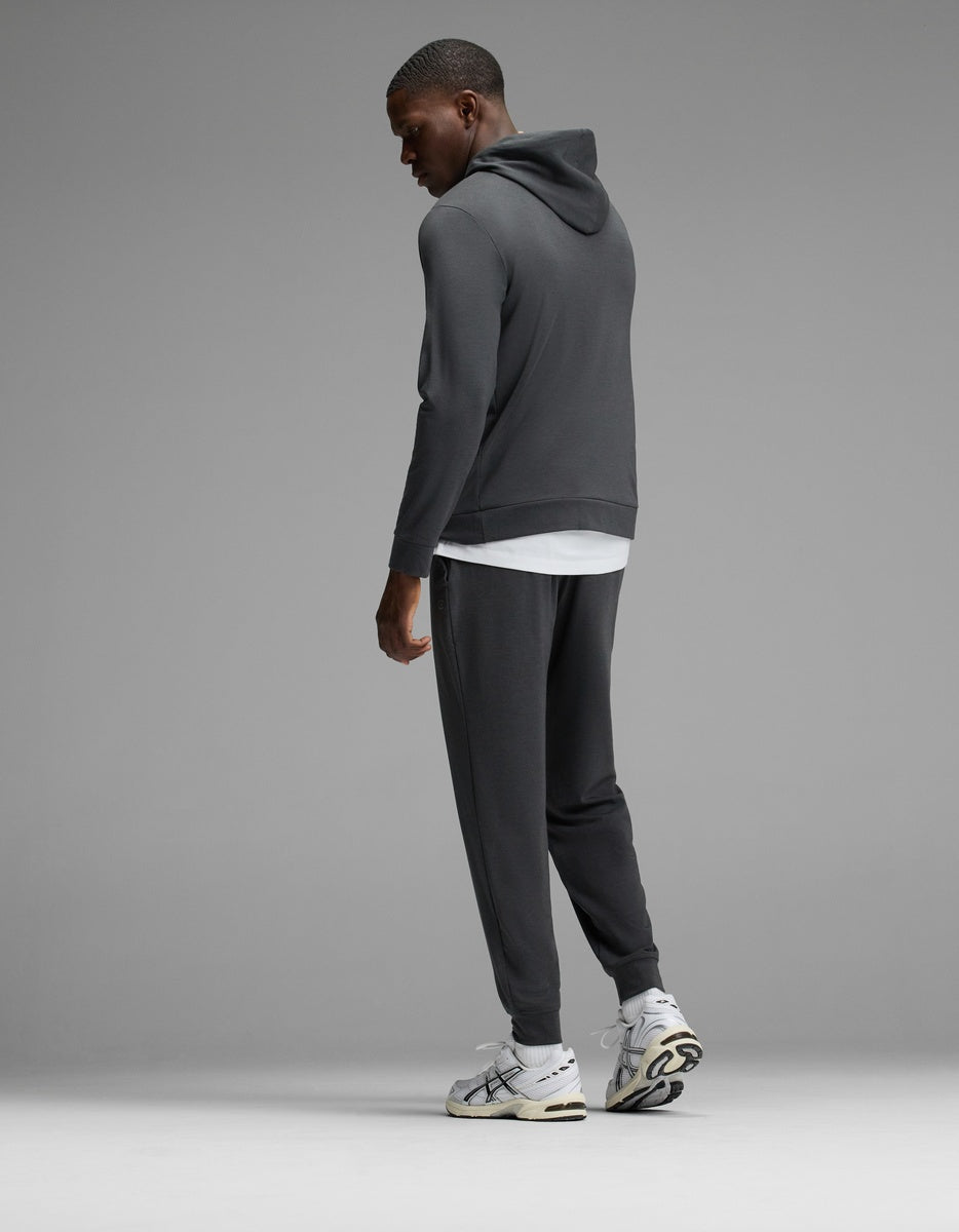 A man wearing the Cozy Earth Men’s Brushed Bamboo Hoodie & Jogger Set in dark gray, paired with a white t-shirt and white sneakers, stands facing away and looks over his shoulder against a plain gray background.