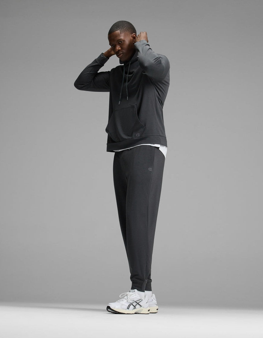A man wearing the Cozy Earth Men’s Brushed Bamboo Hoodie & Jogger Set stands adjusting his hood, paired with white sneakers. The background is plain gray.