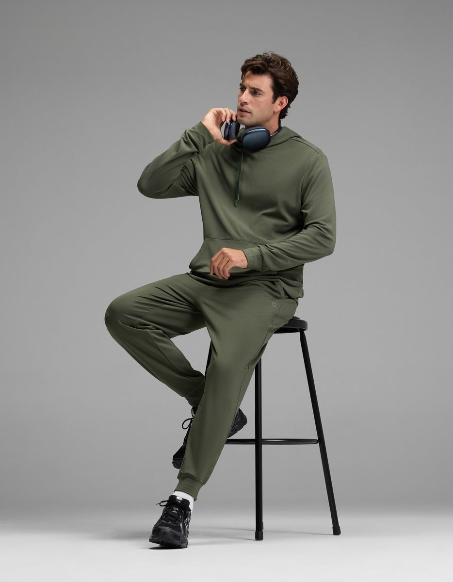 A man wearing the Cozy Earth Men’s Brushed Bamboo Hoodie & Jogger Set in olive green sits on a black stool, holding a phone to his ear with headphones resting around his neck, against a plain gray background.