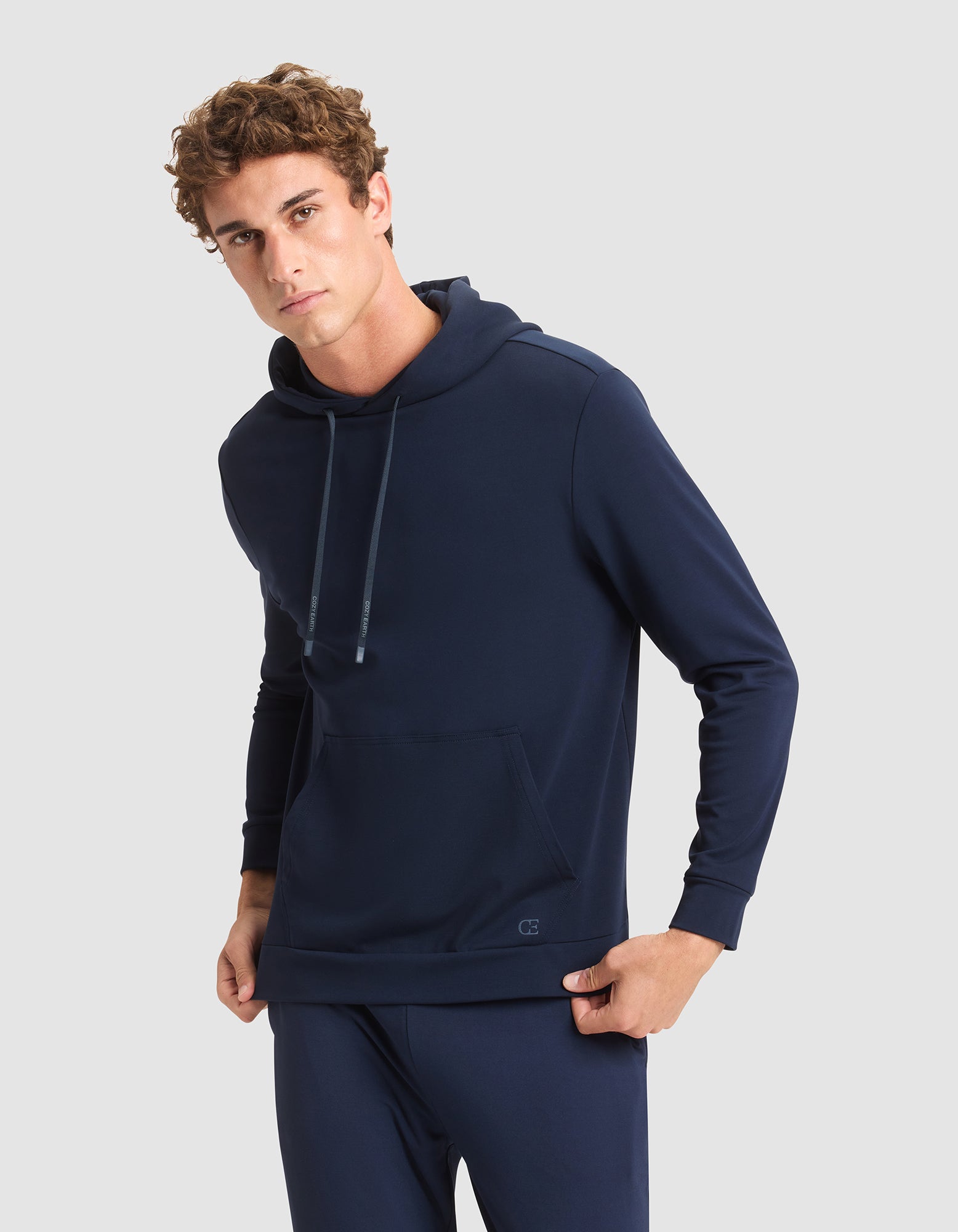 A young man wears the Cozy Earth Men's Brushed Bamboo Hoodie in navy blue, paired with matching pants, standing against a plain light gray background and looking at the camera with a neutral expression. 