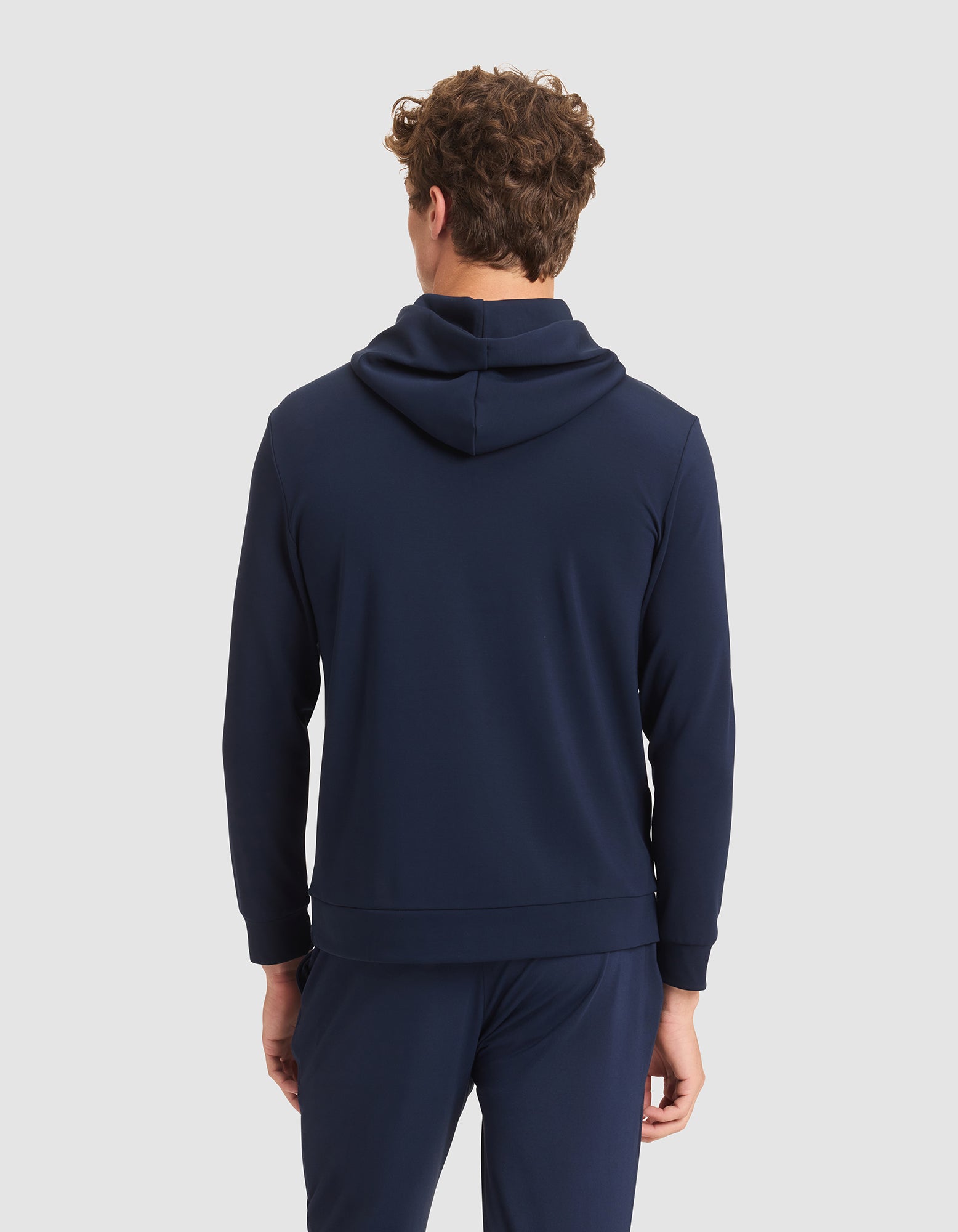 A person with curly brown hair is shown from behind, wearing the Cozy Earth Men's Brushed Bamboo Hoodie in navy and matching pants against a plain light gray background. 
