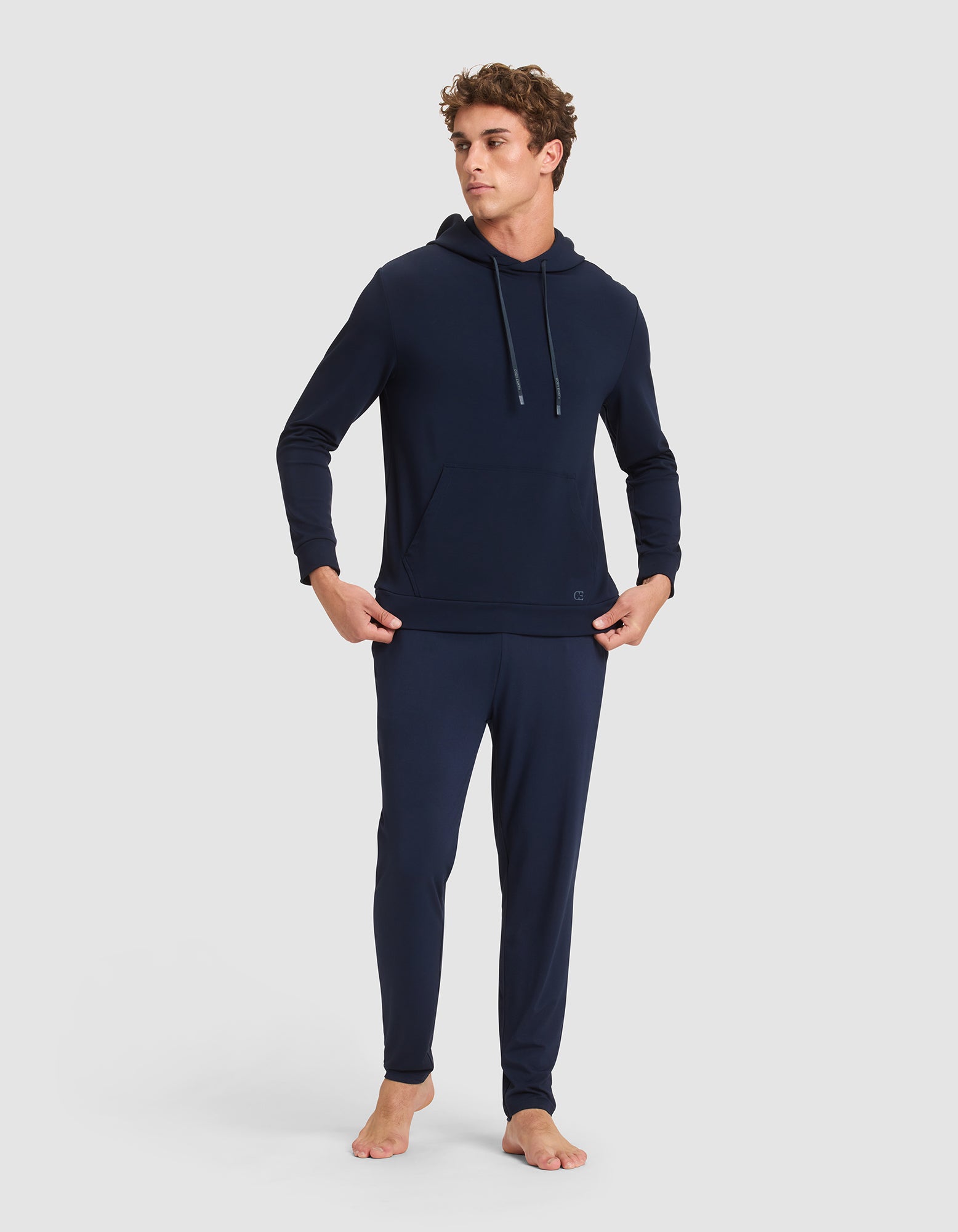 A man stands barefoot in the Cozy Earth Men's Brushed Bamboo Hoodie, featuring a front pocket and a navy hue, paired with matching jogger pants against a plain light gray background.  