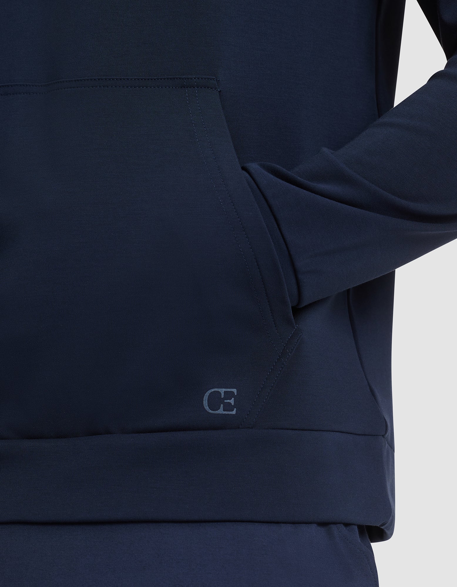 A person wears the Cozy Earth Men's Brushed Bamboo Hoodie in dark blue, with one hand in the front pocket. A small "CE" logo is visible on the hem. The background is plain and light colored. 