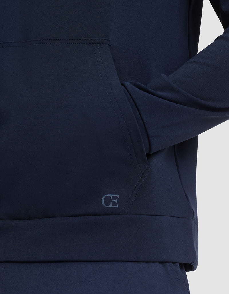 A person wears the Cozy Earth Men's Brushed Bamboo Hoodie in dark blue, with one hand in the front pocket. A small "CE" logo is visible on the hem. The background is plain and light colored.