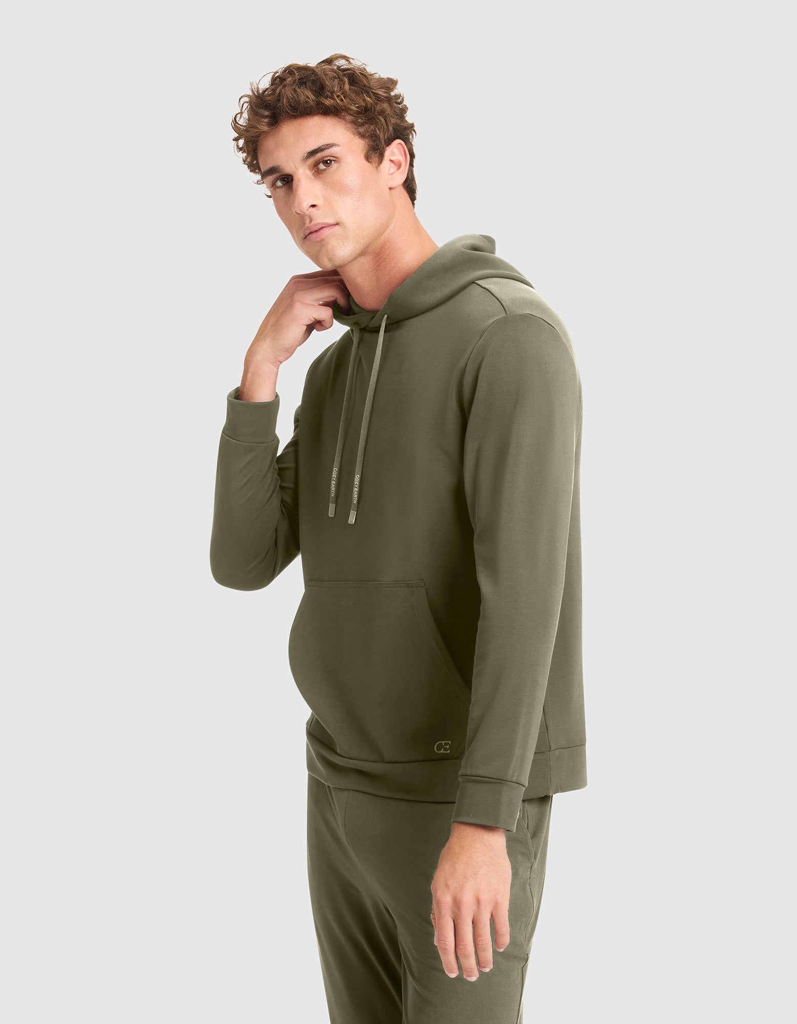 A young man with curly hair models the Cozy Earth Men's Brushed Bamboo Hoodie in olive green, paired with matching pants, standing against a plain light gray background and looking at the camera while touching his neck. 
