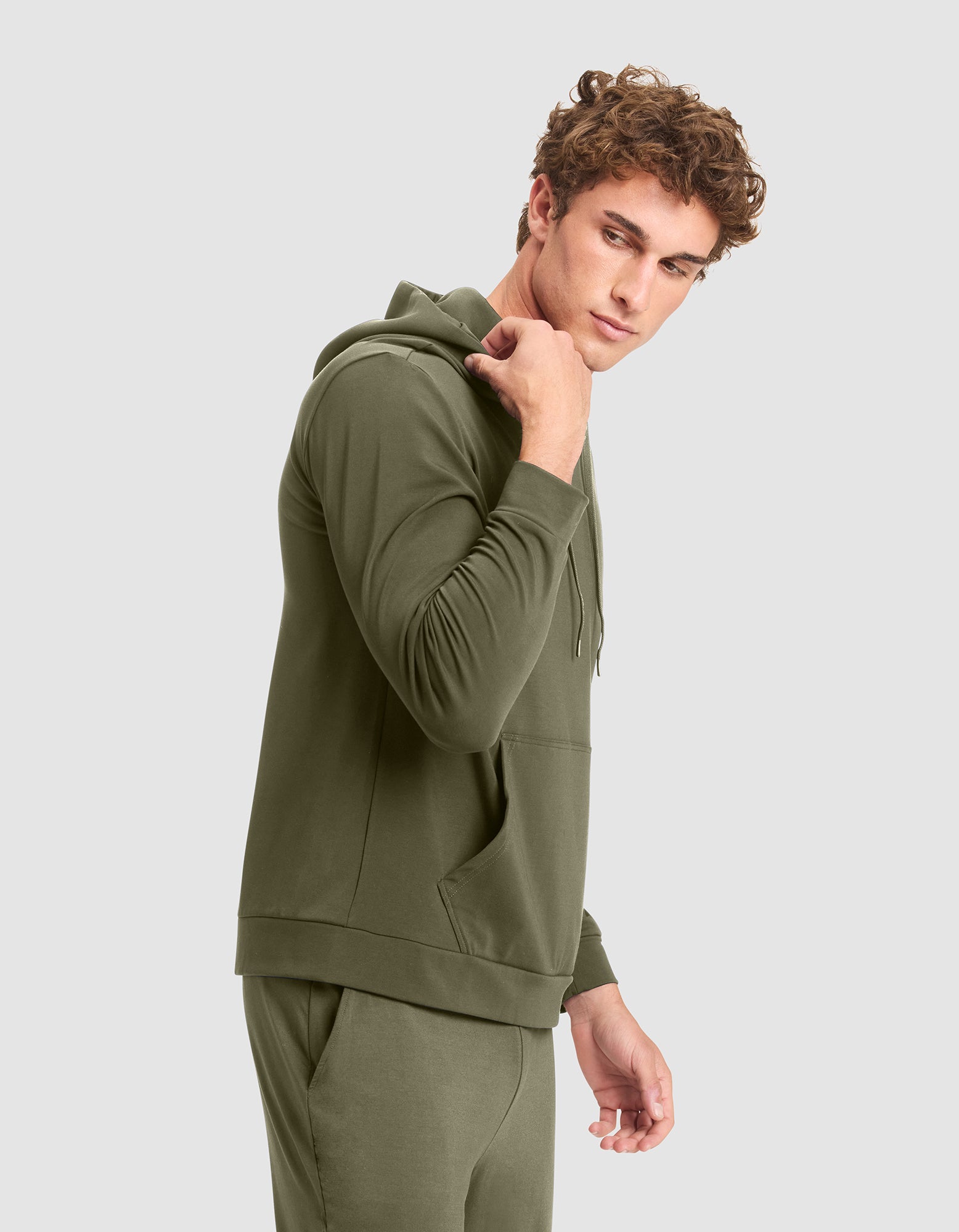 A young man with curly hair models the Cozy Earth Men's Brushed Bamboo Hoodie in olive green, paired with matching joggers, standing sideways and glancing over his shoulder against a plain light gray background. 