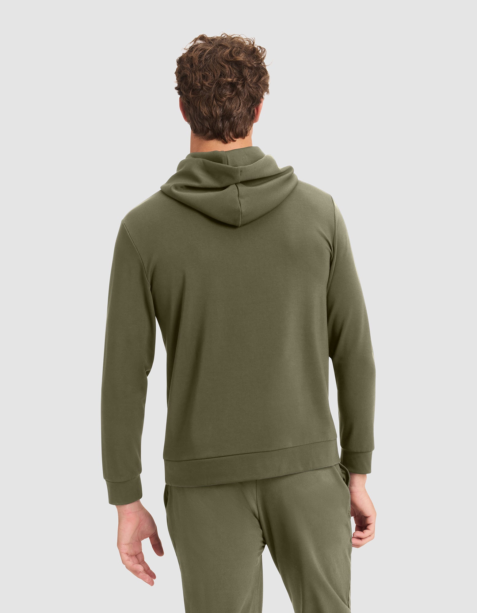 A man faces away, wearing the Cozy Earth Men's Brushed Bamboo Hoodie in olive green with matching sweatpants, his left hand relaxed at his side. The background is light gray. 