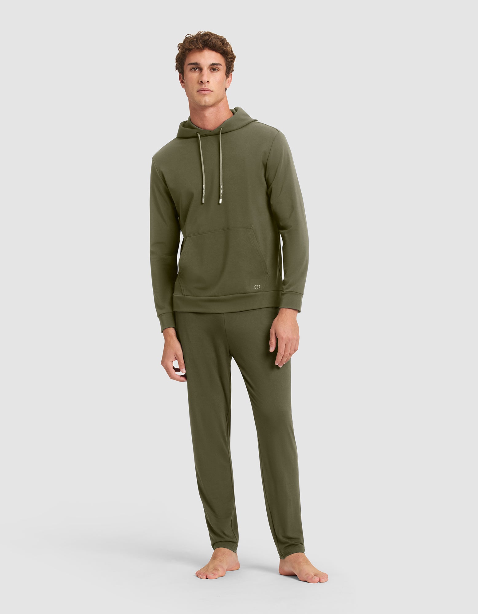 A young man stands barefoot in Cozy Earth's Men's Brushed Bamboo Hoodie—an olive green sweatshirt with drawstrings and matching lounge pants—set against a plain light gray background. 