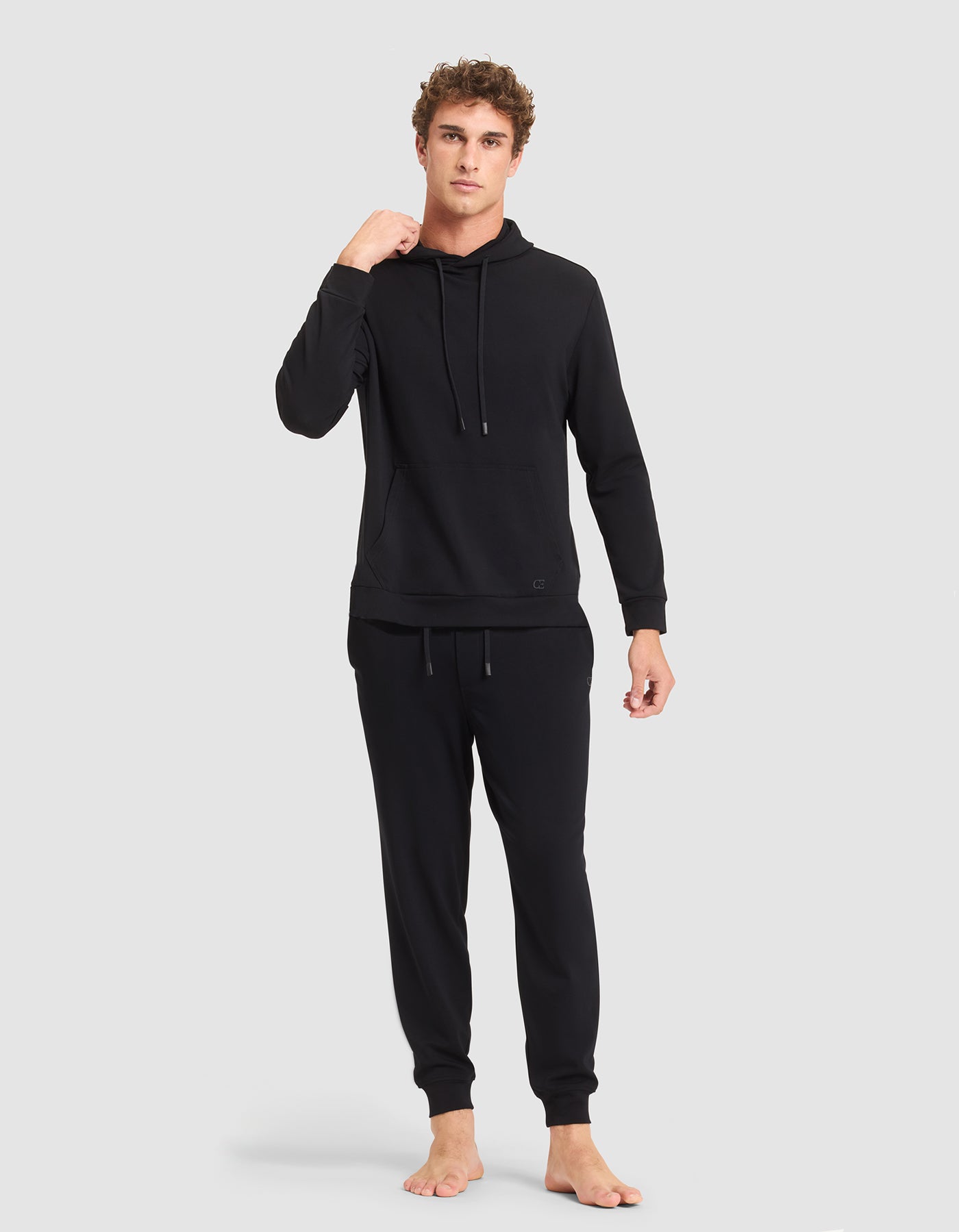 A person stands barefoot against a plain background, wearing the Cozy Earth Men's Brushed Bamboo Hoodie & Jogger Set in black, with their left hand near their neck and looking straight ahead with a neutral expression. 