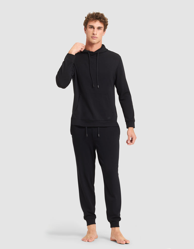 A person stands barefoot against a plain background, wearing the Cozy Earth Men's Brushed Bamboo Hoodie & Jogger Set in black, with their left hand near their neck and looking straight ahead with a neutral expression.