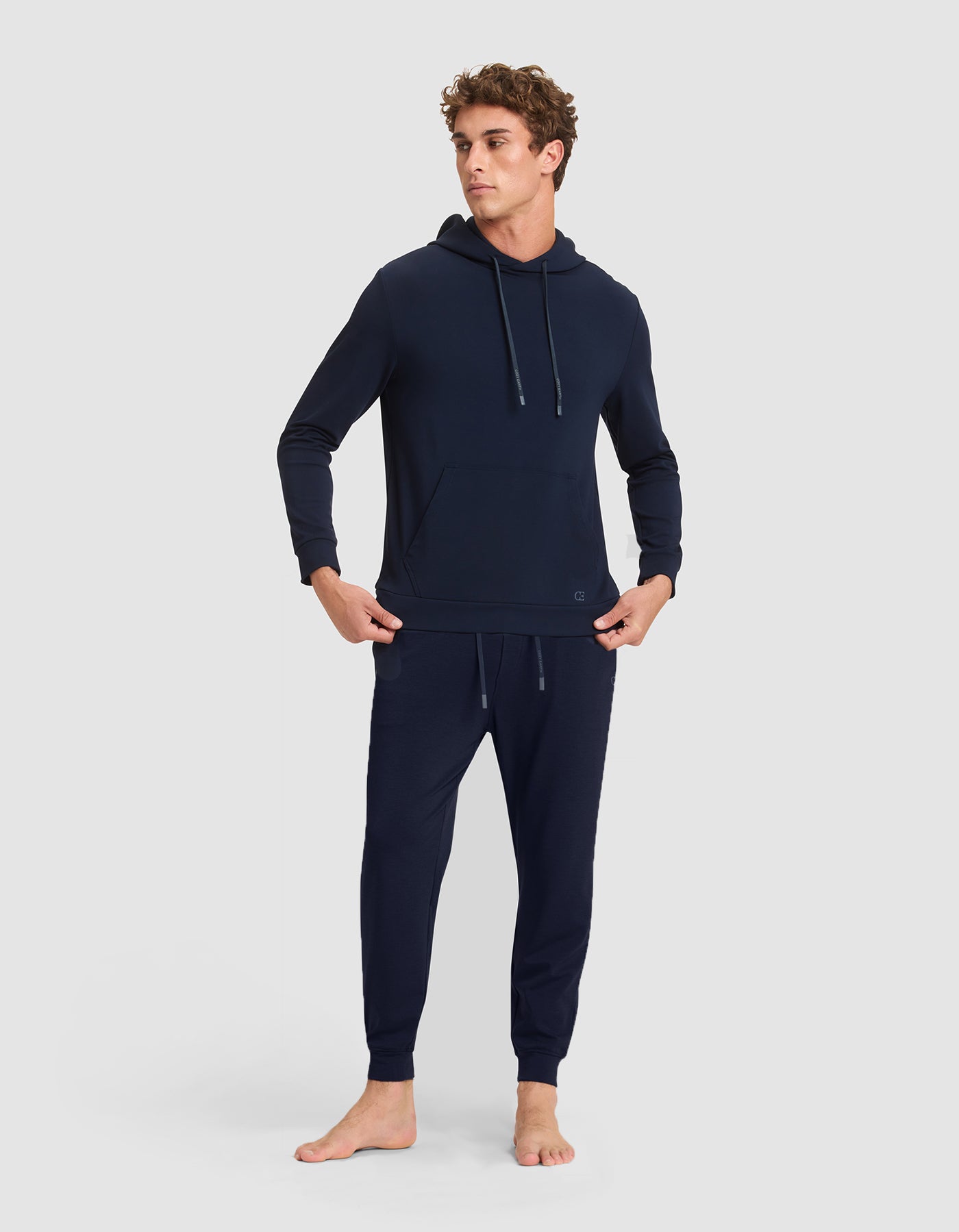A young man stands barefoot against a light gray background, wearing the Cozy Earth Men's Brushed Bamboo Hoodie & Jogger Set with his hands in his pockets. 