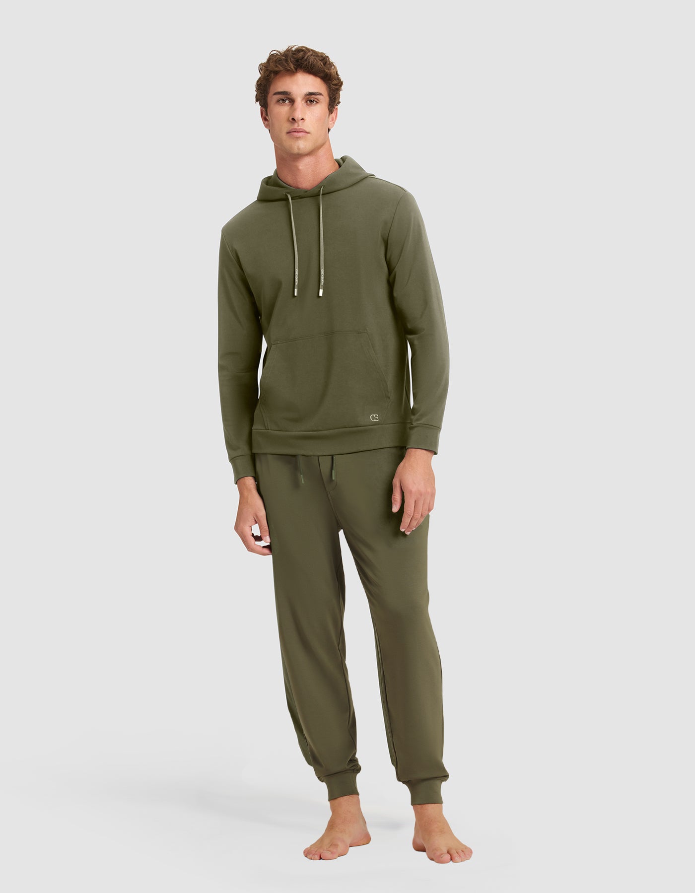 A young man stands barefoot against a gray background, wearing the Cozy Earth Men's Brushed Bamboo Hoodie & Jogger Set in olive green, featuring a hood with drawstrings and matching jogger pants. 