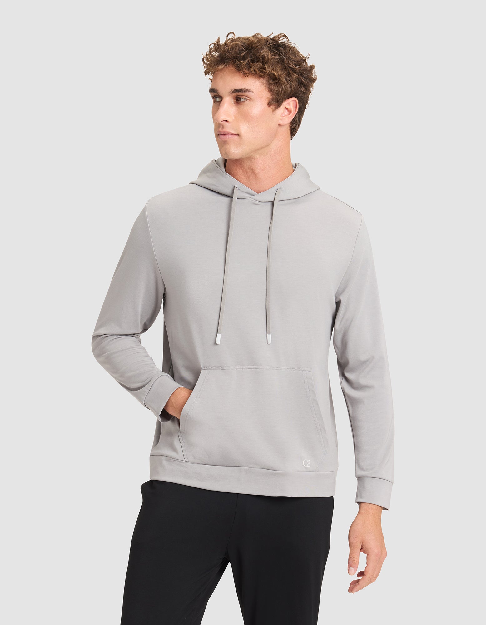 A young man with curly hair models the Cozy Earth Men's Brushed Bamboo Hoodie in light gray, paired with black pants. He stands against a plain light background, one hand in the hoodie pocket, looking to the side. 