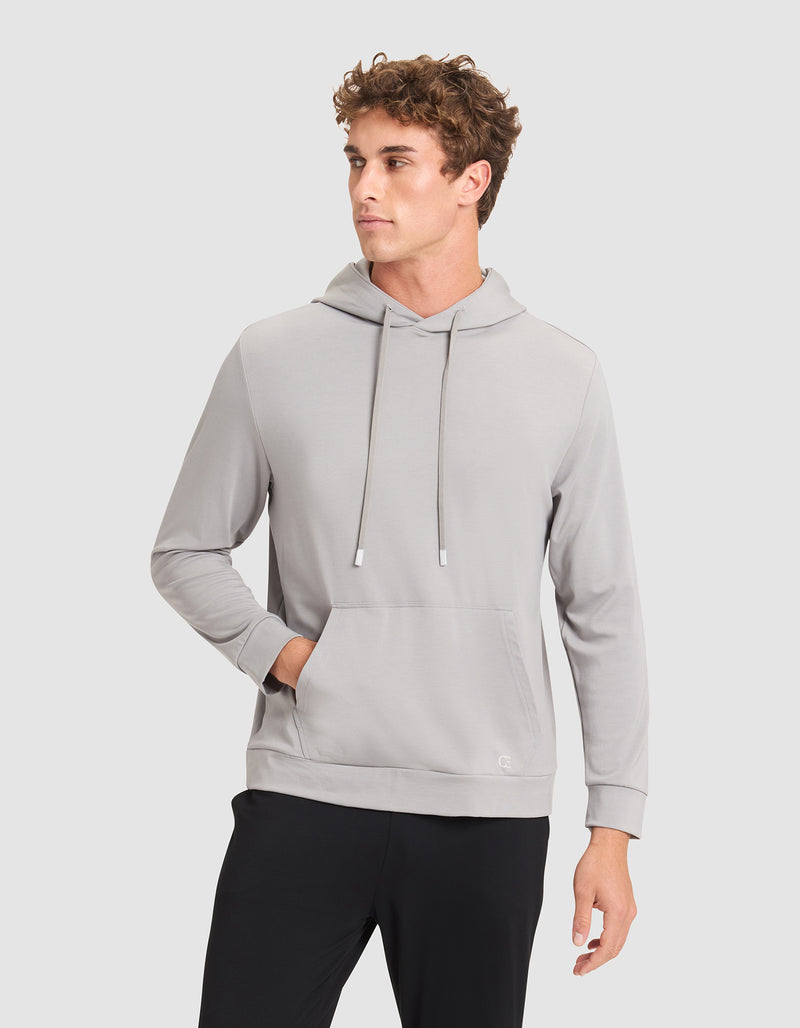 A young man with curly hair models the Cozy Earth Men's Brushed Bamboo Hoodie in light gray, paired with black pants. He stands against a plain light background, one hand in the hoodie pocket, looking to the side.