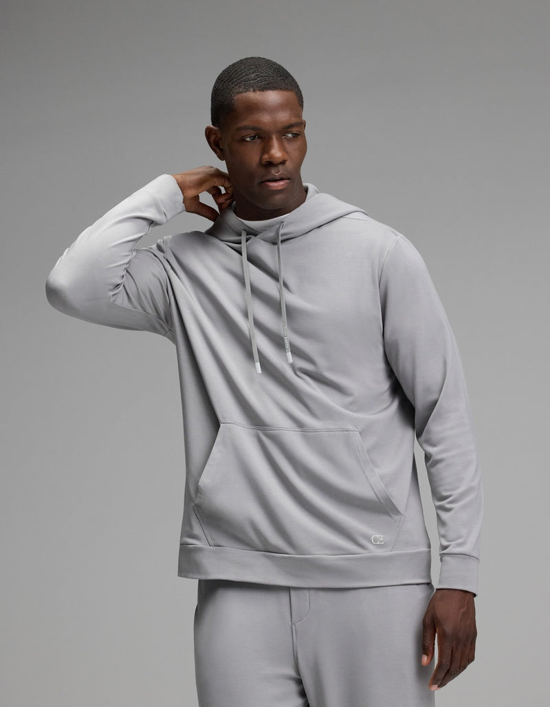 A man wearing the Cozy Earth Men’s Brushed Bamboo Hoodie and matching pants stands against a plain gray background, with one hand touching the back of his neck and the other hanging by his side.
