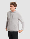 A young man stands against a plain light gray background, wearing Cozy Earth's Men's Brushed Bamboo Hoodie in light gray and black pants, looking to the side with his hands pulling at the hem of the hoodie. |Color:Stone