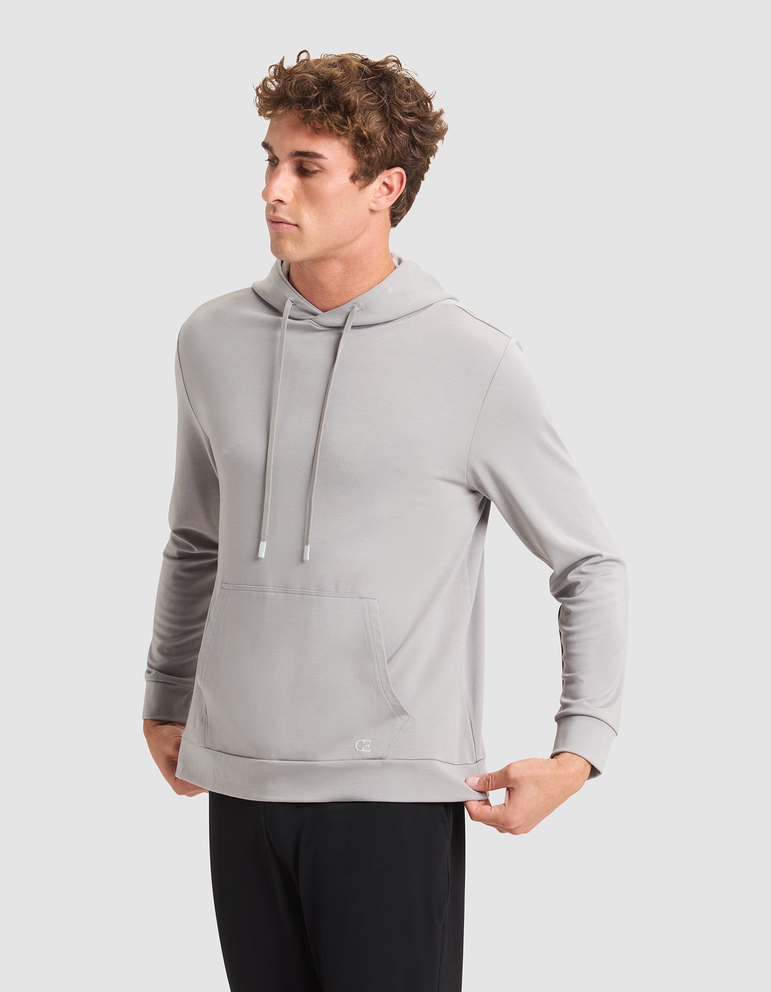 A young man stands against a plain light gray background, wearing Cozy Earth's Men's Brushed Bamboo Hoodie in light gray and black pants, looking to the side with his hands pulling at the hem of the hoodie. 