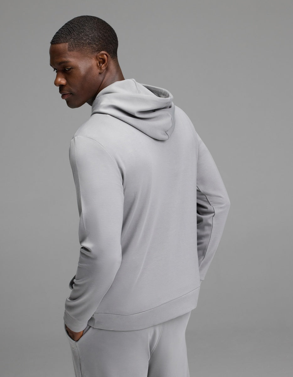 A person wearing the Cozy Earth Men’s Brushed Bamboo Hoodie in light gray, paired with matching pants, stands with hands in pockets and faces slightly away against a plain gray background.