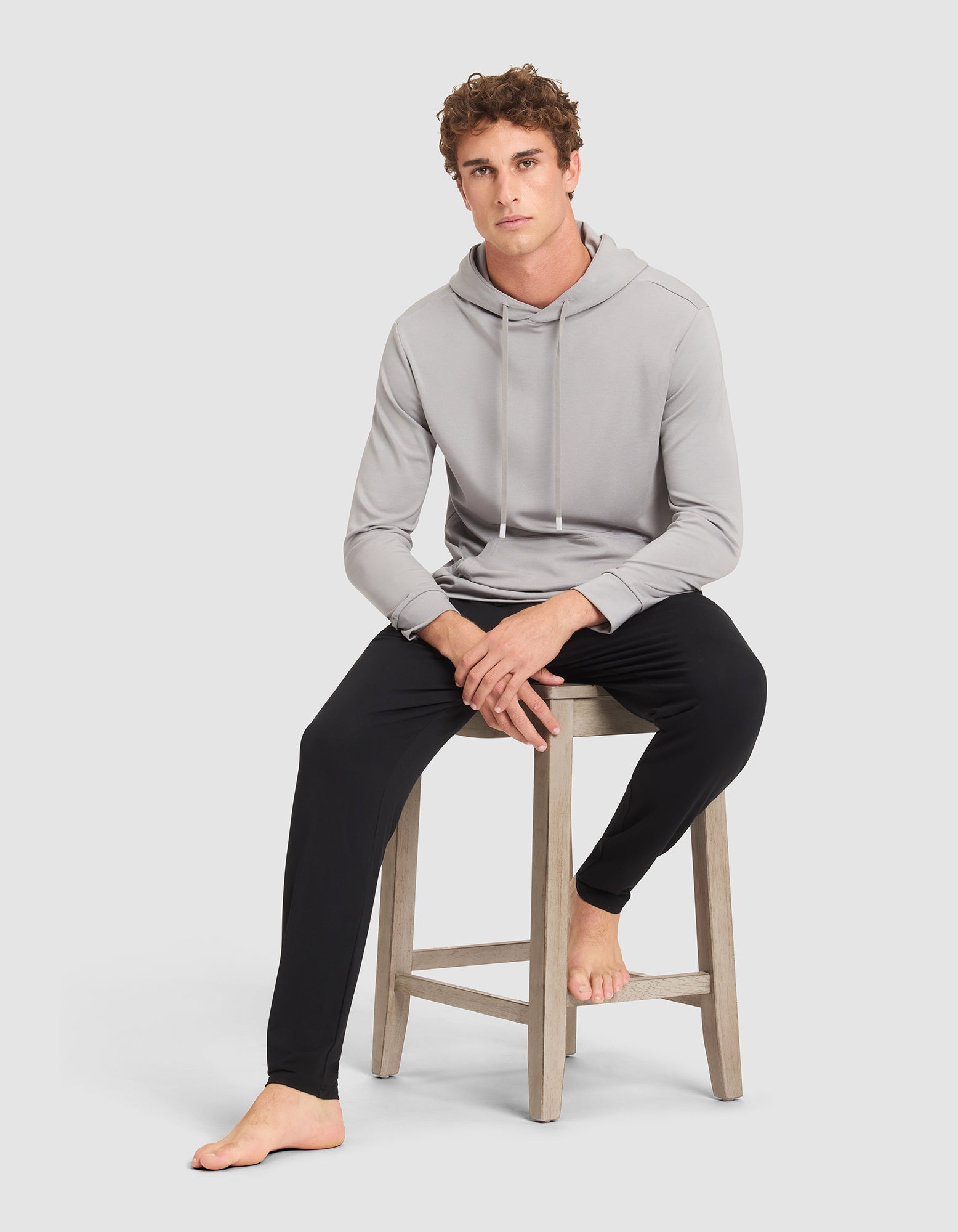 A young man with curly hair models the Cozy Earth Men's Brushed Bamboo Hoodie in light gray and black pants, sitting barefoot on a wooden stool against a plain light gray background. 