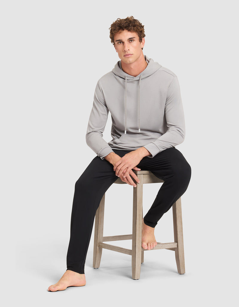 A young man with curly hair models the Cozy Earth Men's Brushed Bamboo Hoodie in light gray and black pants, sitting barefoot on a wooden stool against a plain light gray background.