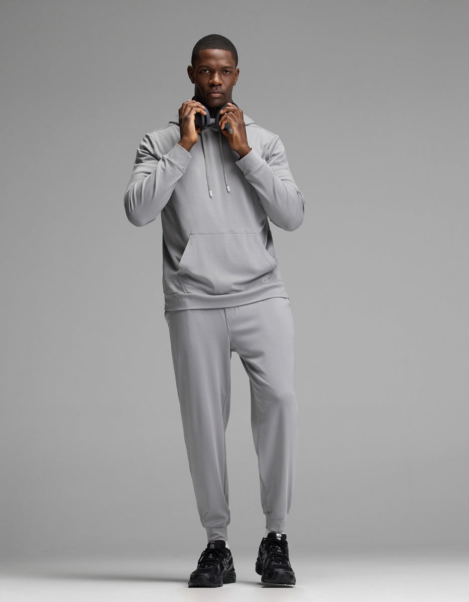 A person stands facing forward against a plain gray background, wearing Cozy Earth’s Men’s Brushed Bamboo Hoodie in light gray along with matching joggers and black sneakers, adjusting the hood drawstrings.