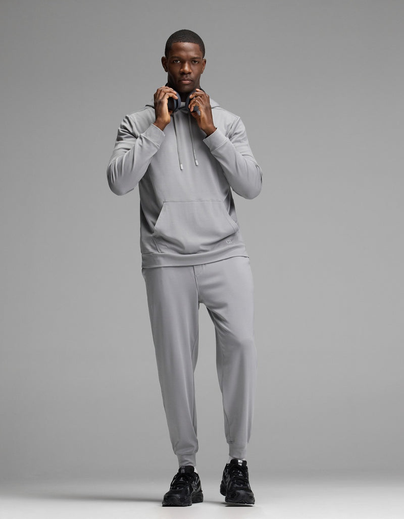 A person stands facing forward against a plain gray background, wearing Cozy Earth’s Men’s Brushed Bamboo Hoodie in light gray along with matching joggers and black sneakers, adjusting the hood drawstrings.