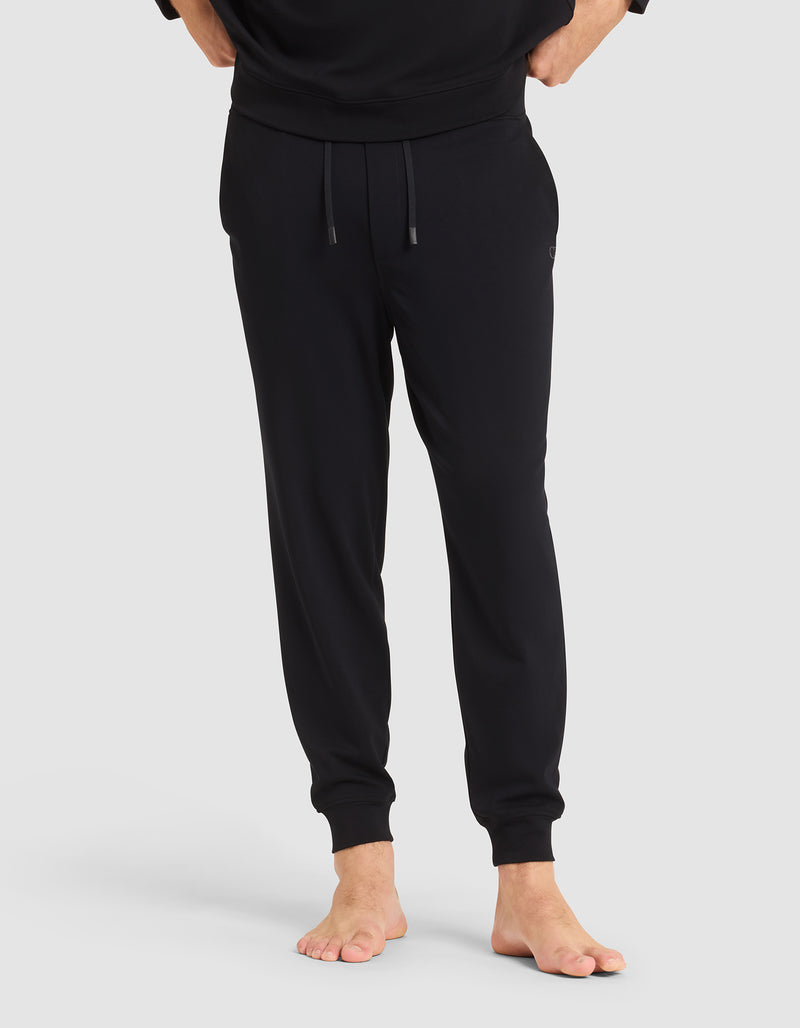 A person stands barefoot in Cozy Earth's Men's Brushed Bamboo Jogger Pant—black, with an elastic waistband and drawstrings, hands in pockets—against a plain light gray background.