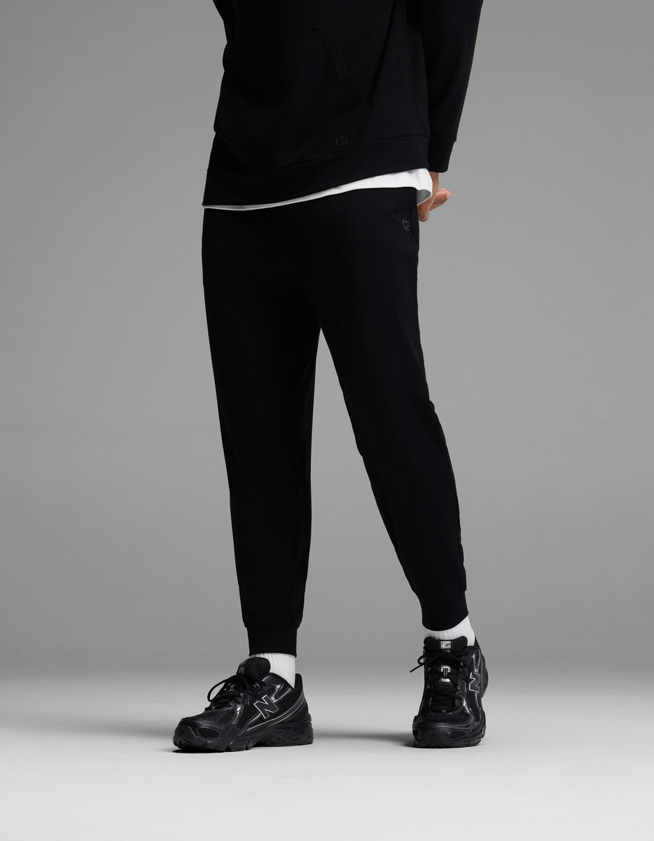 A person is shown from the shoulders down, standing against a gray background and wearing Cozy Earth’s Men’s Brushed Bamboo Joggers in black, paired with a black hoodie, white T-shirt, white socks, and black sneakers. The head is not visible in the image.|Color:Black