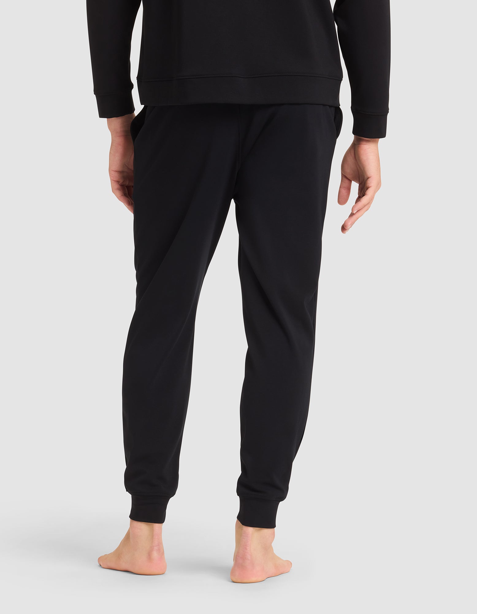 A person models Cozy Earth's Men's Brushed Bamboo Jogger Pant in black, paired with a matching sweatshirt, standing barefoot and facing away from the camera against a light gray background.