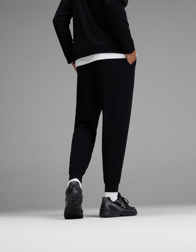 A person stands facing away, wearing black jogger pants, a black jacket, a white shirt, white socks, and black sneakers against a gray background.