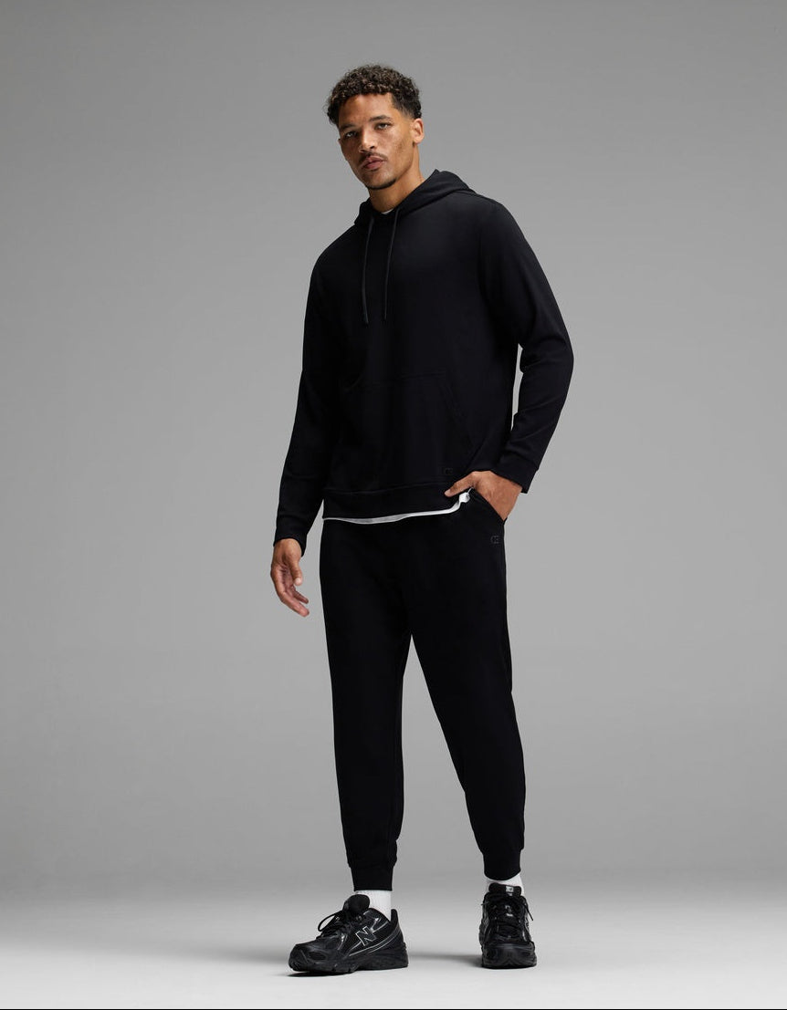 A man stands against a gray background wearing a black hoodie, black jogger pants, and black sneakers. He has short curly hair and is looking at the camera with a relaxed posture.
