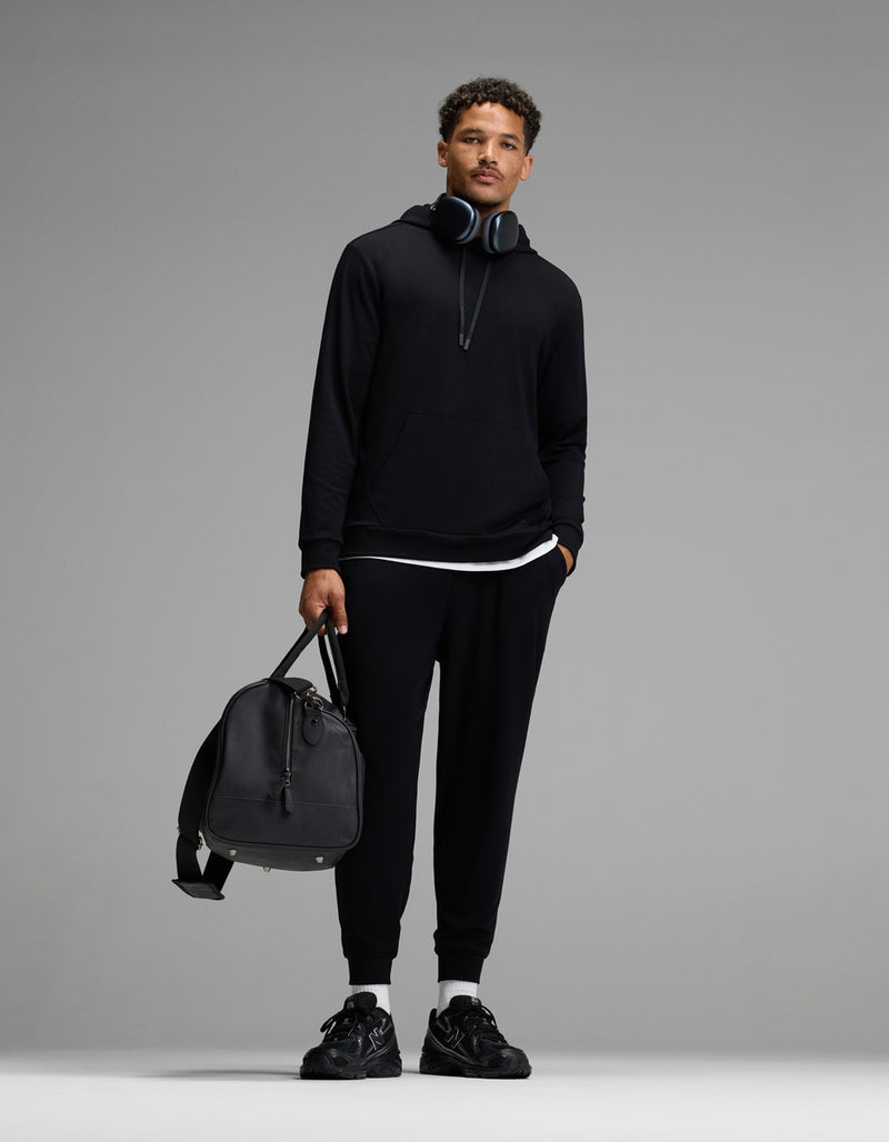 A man stands against a plain gray background wearing Cozy Earth’s Men’s Brushed Bamboo Joggers and a black hoodie. He holds a black duffel bag, wears black sneakers, and has headphones around his neck.